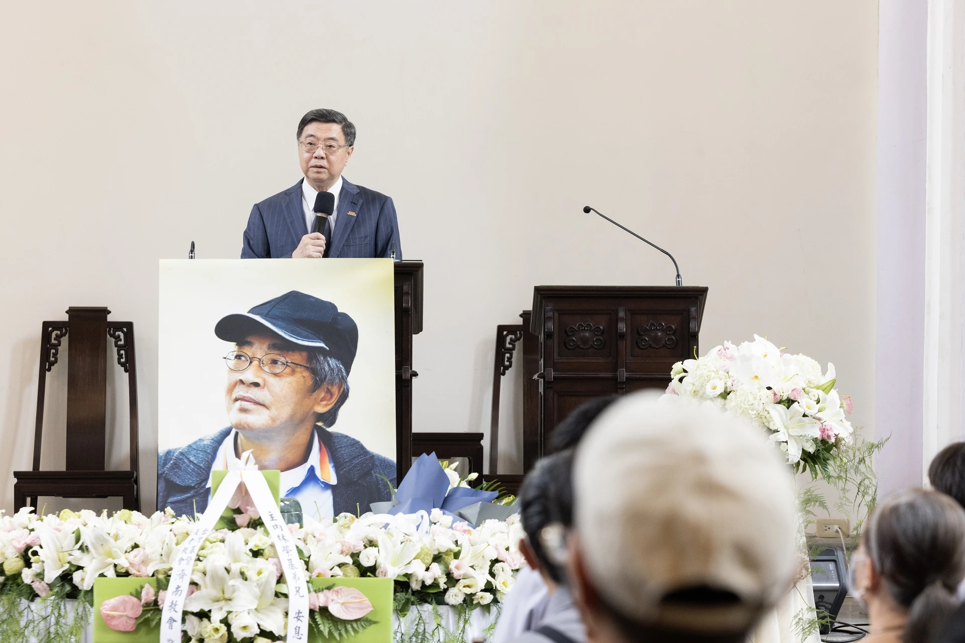 行政院長卓榮泰站在講臺前手持麥克風發言，講臺下方擺放著林榮基的遺照與白色花籃裝飾。
