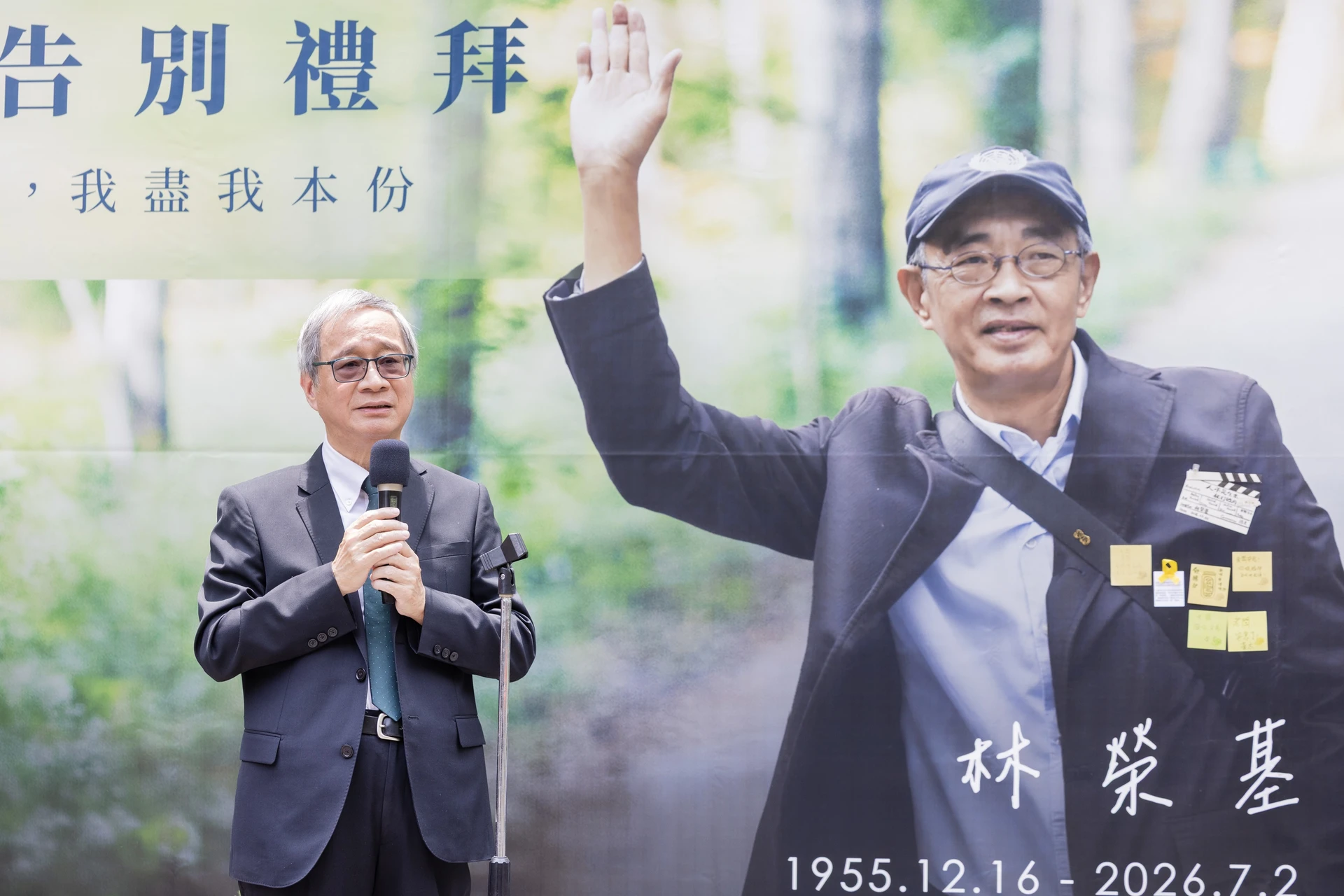 文化部長李遠身穿西裝、打領帶，手持麥克風在講臺前發言，背景為林榮基舉手揮別的巨大海報與生卒年。