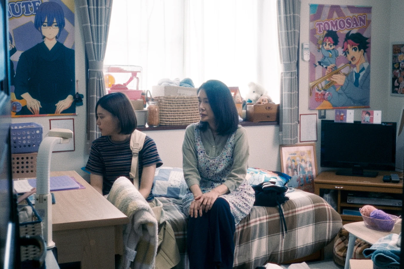 電影劇照中，杉咲花坐在房間書桌旁，身旁坐著資深女演員，背景充滿動漫周邊與海報。