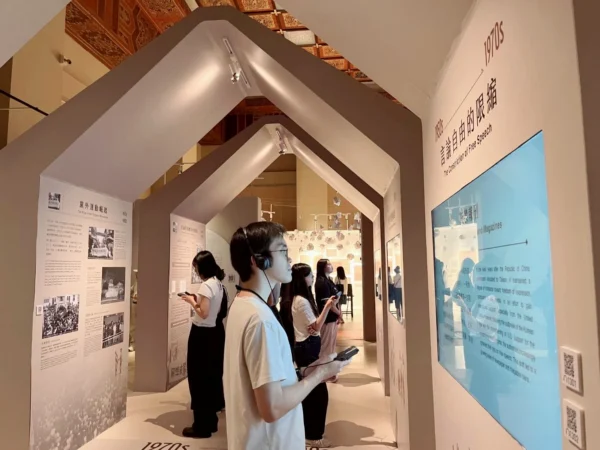 一名年輕男性在展場中戴著耳機聆聽語音導覽，前方為展覽牆面與多媒體螢幕。