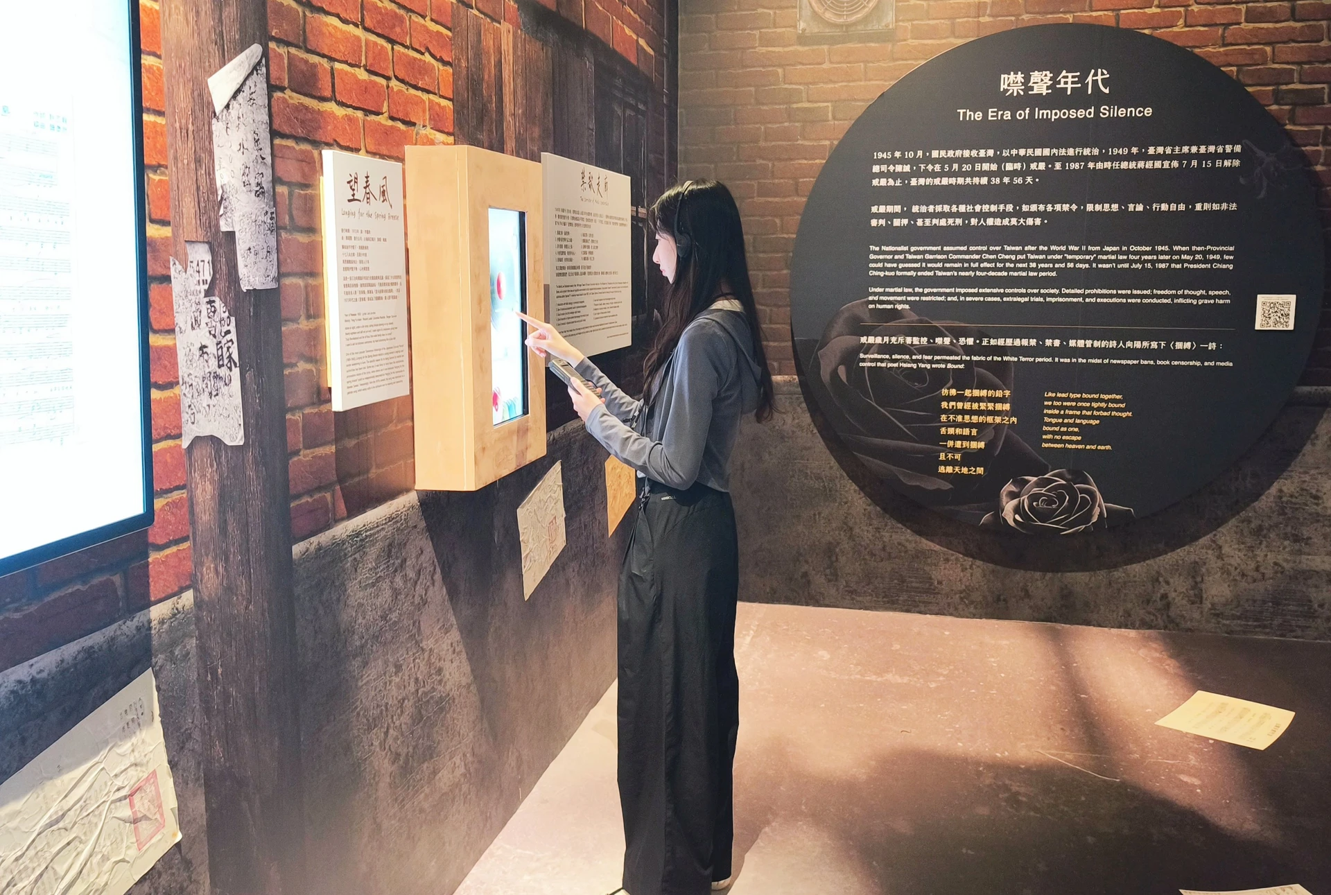 一名女性觀眾在展場互動裝置前，一邊戴著耳機聆聽語音導覽，一邊操作觸控螢幕。